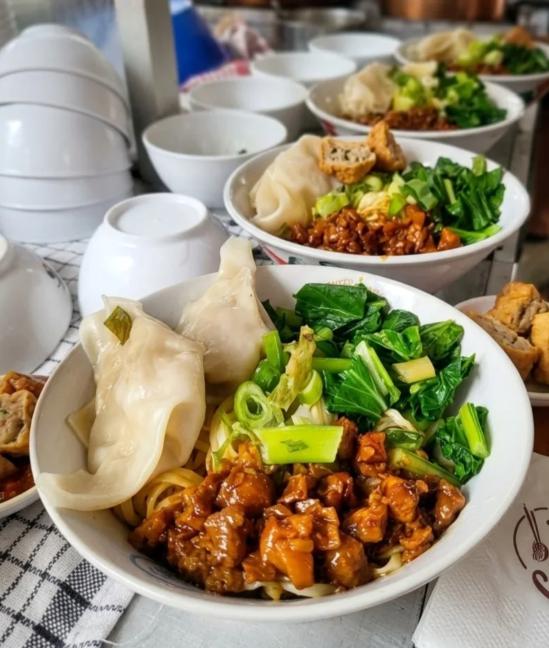Semangkuk bakmi dengan pangsit basah, sayur hijau dan topping saus kunpow