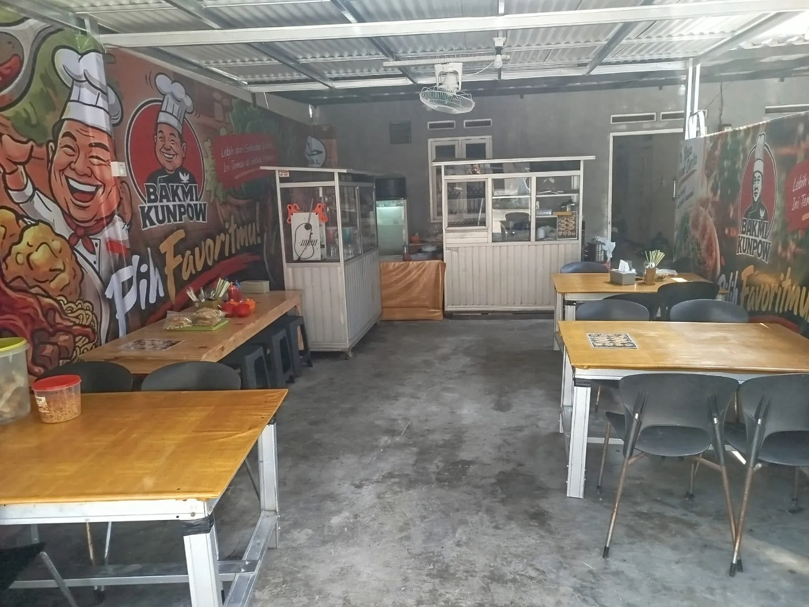 Area makan di dalam warung dengan meja kayu dan mural koki